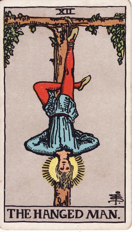 The Hanged Man tarot card from the Rider-Waite-Smith deck: a man suspended by one ankle from a living T-shaped tree, a halo of light around his head.