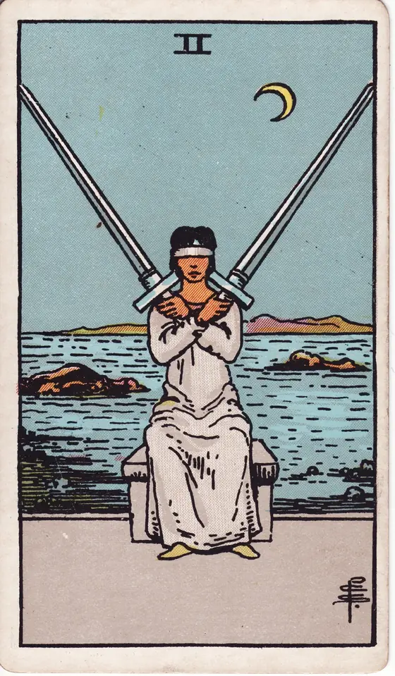 Two of Swords tarot card from the Rider-Waite-Smith deck: a blindfolded woman seated at the shore holding two crossed swords.