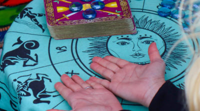 blond lady with both hands on table with palm up. The table cloth slight blue with the sun in the middle and the zodiac symbols around it. Tarot cards are also on the table.